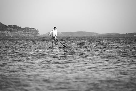 grayscale photo of man holding paddle on body of water thumbnail