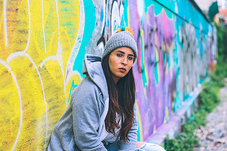 girl in gray jacket and bobble hat sitting on concrete wall with bench and painted graffiti at day time thumbnail