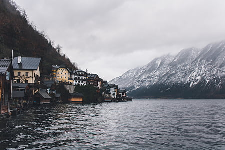 buildings near body of water and snow covered mountain under gray sky during daytime thumbnail