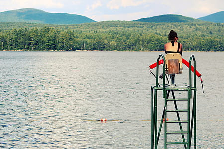 woman sitting near calm body of water across green leaf tree and mountain under blue sky thumbnail