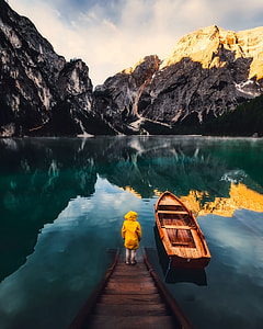 woman in yellow hooded jacket on the brown wooden dock near the row boat on the lake overlooking the snow capped mountain range under the cloudy daytime skies thumbnail