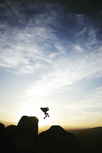 silhouette of person jumping on rock formation under white cloud blue skies during sunrise thumbnail