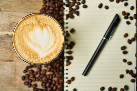 coffee cup filled with cappuccino and black gel pen on white notebook thumbnail