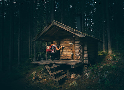 woman in grey shirt with red backpack about to enter a small brown house surrounded with trees thumbnail