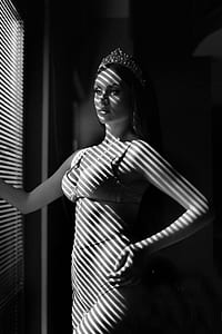 grayscale photo of woman wearing crown standing in front of window blinds thumbnail