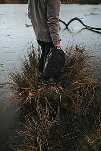 person standing on brown grass holding backpack surrounded by water thumbnail