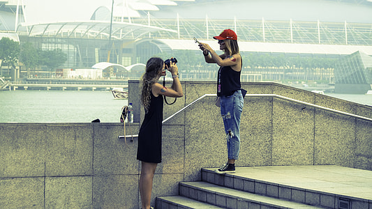two women's taking picture each other on gray concrete pathway beside river thumbnail