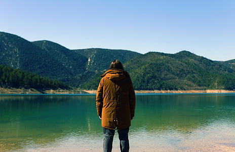 woman standing in front of body of water near mountains under blue sky during daytime thumbnail