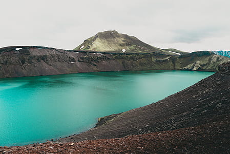 green calm body of water surrounded by hills under white sky thumbnail