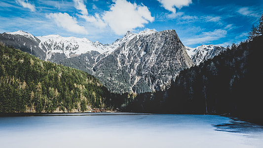 lake surrounded by trees near snow-covered mountain during daytime thumbnail