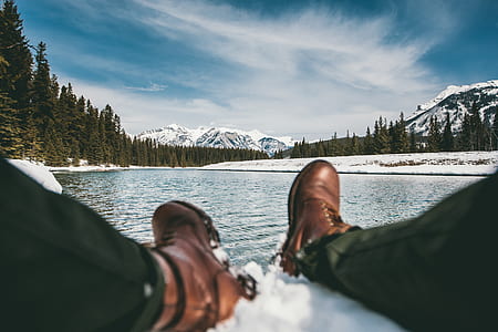 person sitting on snow near calm body of water surrounded by trees and mountain under white cloudy sky during daytime thumbnail