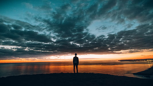 silhouette photo of man standing on high ground looking at the body of water thumbnail