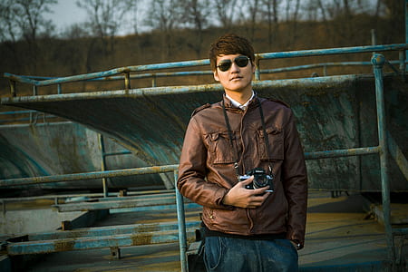 man wearing brown leather zip-up jacket holding black DSLR camera thumbnail