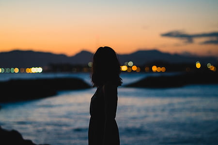 silhouette photo of a woman standing near body of water under cloudy sky during golden hour thumbnail