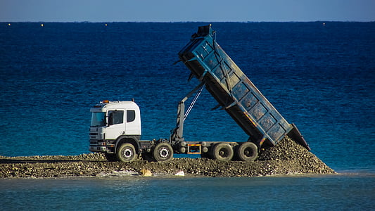 white and blue dump truck dumping gravel near body of water thumbnail