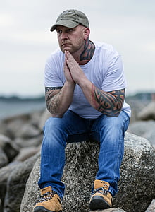 tattooed man wearing gray baseball cap, white crew-neck t-shirt, and blue denim jeans sitting on gray boulder under gray clouds thumbnail