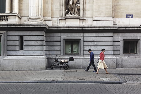 woman wearing dress passed by walking man beside street side with parked motorcycled beside concrete building thumbnail