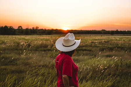 person in red shirt wearing white cowboy hat standing on grass field during golden hour thumbnail