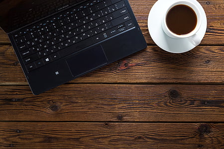 black laptop computer beside white ceramic teacup with saucer on brown wooden surface thumbnail