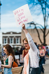 woman in white cardigan and pink shirt walking on street while holding paper board with text thumbnail