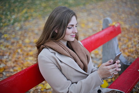 Young Woman Messaging On Her Mobile Phone While Sitting On Red Bench In Autumn Park thumbnail