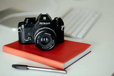 black Nikon point-and-shoot camera on brown notebook next to ballpoint pen thumbnail