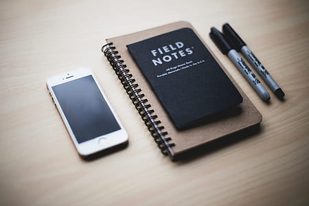 gold iPhone 5s beside notebook and two markers on brown surface thumbnail
