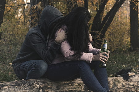 woman in pink long-sleeved shirt holding green glass bottle in front of person in black hooded jacket surrounded with trees thumbnail