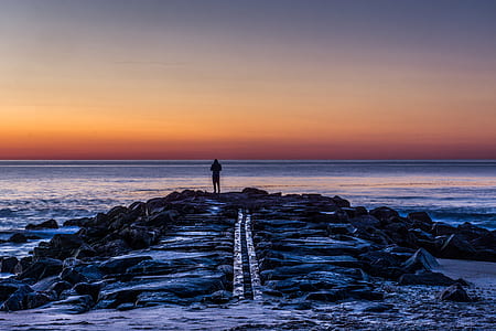 person standing on rock formation near body of water during golden hour thumbnail