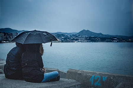 candid photography of man and woman under umbrella facing body of water under blue calm sky thumbnail