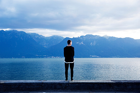 man in black sweater facing the alps across the calm sea during daytime thumbnail