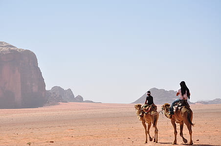 two men riding camels walking on desert with view of brown rock formations during daytime photo thumbnail