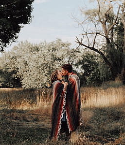 couple wrapped in blanket kissing in the middle of grass field and trees thumbnail