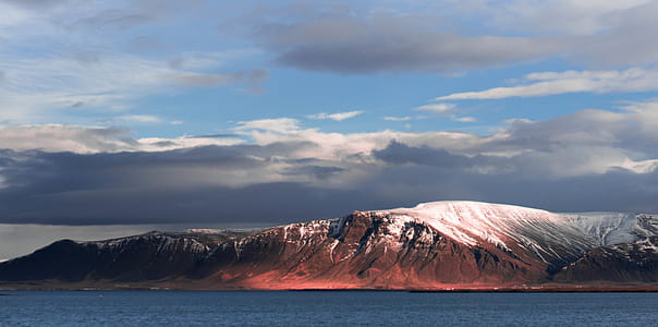 brown rocky mountain beside body of water under dark clouds thumbnail