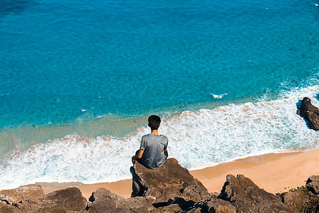 man wearing gray shirt sits on cliff in front of sea during daytime thumbnail