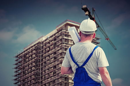man in white hard hat, gray crew-neck t-shirt and blue jumpsuit holding paper watching building thumbnail