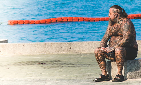Man With Tattoos Sitting on Gray Concrete Floor Near Body of Water thumbnail