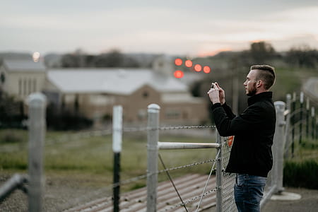 man in black jacket in front of steel fence while holding smartphone during daytime thumbnail
