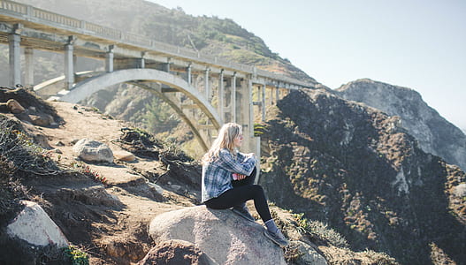 woman in dress shirt sits on grey rock with bridge background thumbnail