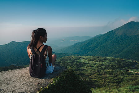 woman sitting on cliff with backpack facing mountain and trees thumbnail