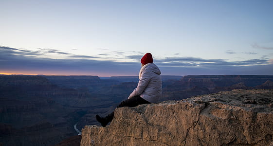 Person Wearing Gray Hooded Jacket and Black Pants Sitting on Mountain Cliff during Sunset thumbnail