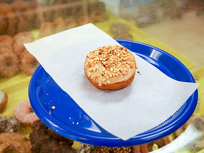 A maple cake donut with peanuts sitting on a blue plate inside a donut shop thumbnail