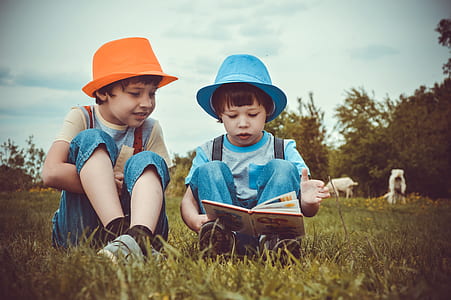 two boys reading book while sitting on green grass field thumbnail
