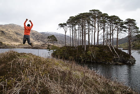 Man in Orange Long-sleeved Shirt Jumping on Lake Near Tall Trees at Daytime thumbnail