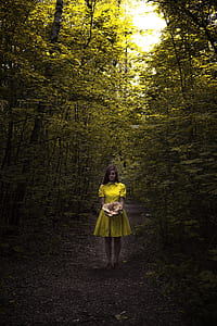 woman wearing yellow button-up collared dress holding brown mushroom while standing between green trees during daytime thumbnail