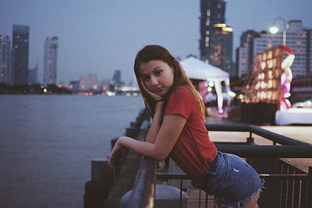 woman leaning on brown metal railings beside body of water with view of city during nighttime photo thumbnail