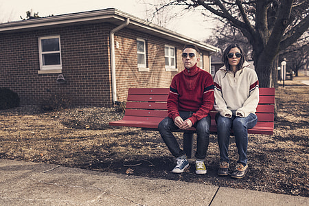 man and woman wearing blue jeans sitting on red wooden outdoor bench plank seat thumbnail