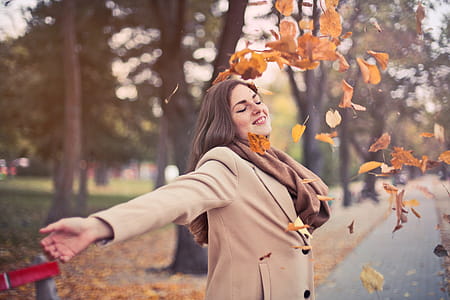 Beautiful young adult woman wearing brown scarf and camel overcoat as playing among falling autumn leaves thumbnail