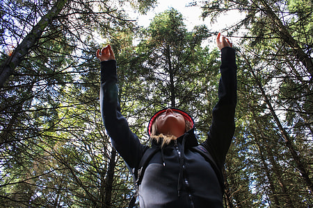 low angle photo of woman wearing black zip-up hooded jacket raising her two hands above with green leaf trees at daytime thumbnail