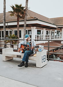 man sits on concrete bench playing guitar near white and brown concrete building during daytime thumbnail